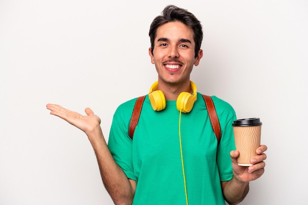 Giovane studente caucasico che beve caffè isolato su sfondo bianco che mostra uno spazio di copia su un palmo e tiene un'altra mano sulla vita.