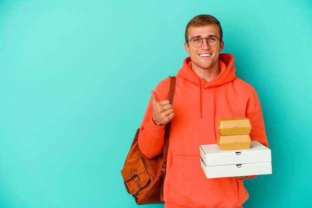 Giovane studente caucasico che tiene hamburger e pizze isolati su blu sorridente e alzando il pollice thumb