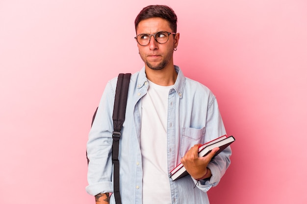 Giovane studente caucasico che tiene in mano libri isolati su sfondo rosa confuso, si sente dubbioso e insicuro.