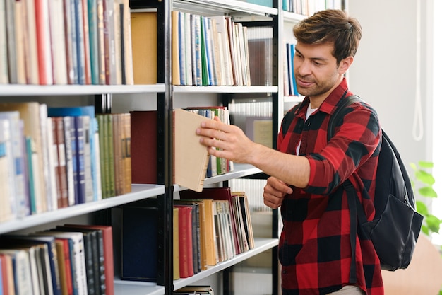 Giovane studente con zaino prendendo uno dei libri dallo scaffale durante la visita alla biblioteca del college