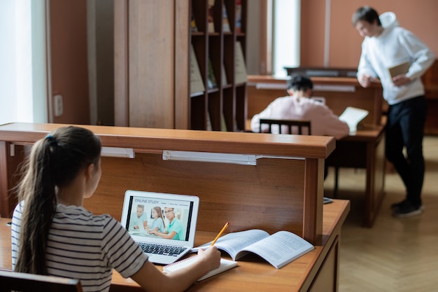 Giovane studente contemporaneo guardando la pagina online del sito web educativo mentre prende appunti nel blocco note in libreria