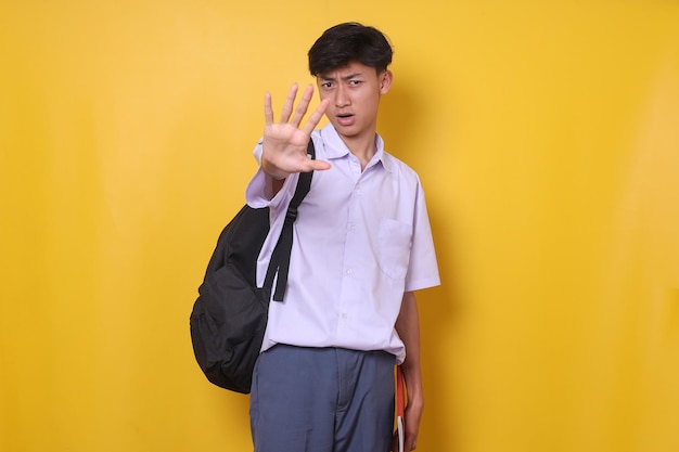 Giovane studente di liceo maschio asiatico in uniforme che fa un gesto di arresto con la mano mentre sta in piedi contro