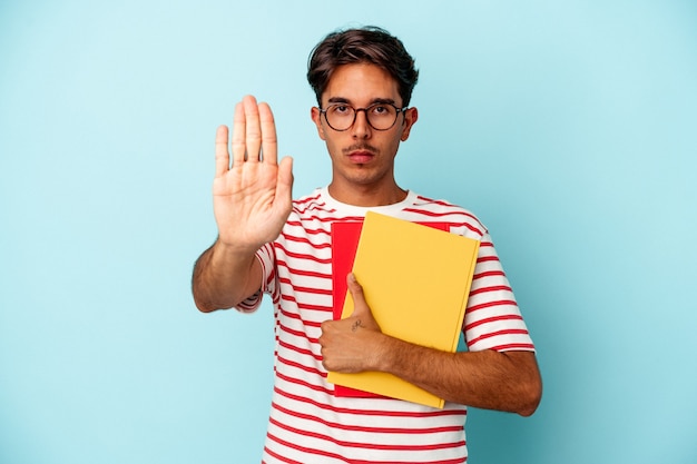 Giovane studente di razza mista che tiene in mano libri isolati su sfondo blu in piedi con la mano tesa che mostra il segnale di stop, impedendoti.