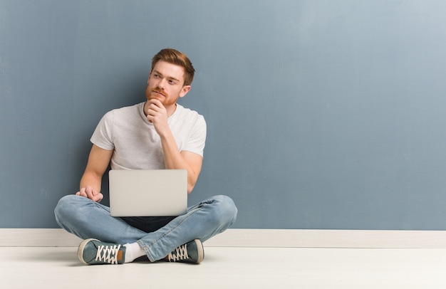 Giovane studente di redhead uomo seduto sul pavimento dubbioso e confuso. Ha in mano un laptop.