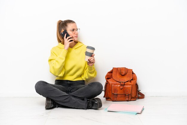 Giovane studente donna caucasica seduta uno il pavimento isolato su sfondo bianco tenendo il caffè da portare via e un cellulare