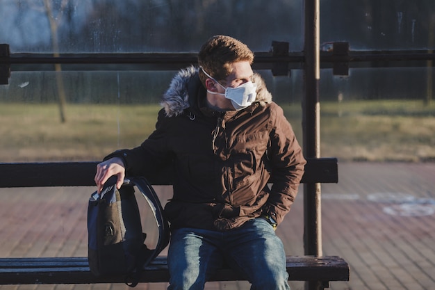 giovane studente è seduto in una maschera a una fermata del trasporto pubblico. Protezione da coronavirus