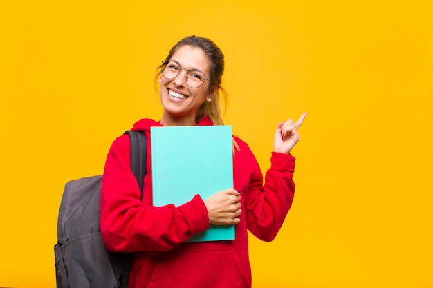 Giovane studente grazioso che sorride felicemente e che indica il lato e verso l'alto con entrambe le mani che mostrano oggetto nello spazio della copia