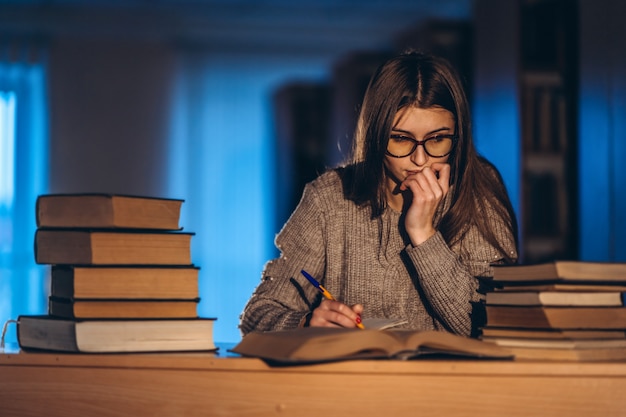 Giovane studente in bicchieri preparando per l'esame. Ragazza di sera si siede a un tavolo in biblioteca con una pila di libri