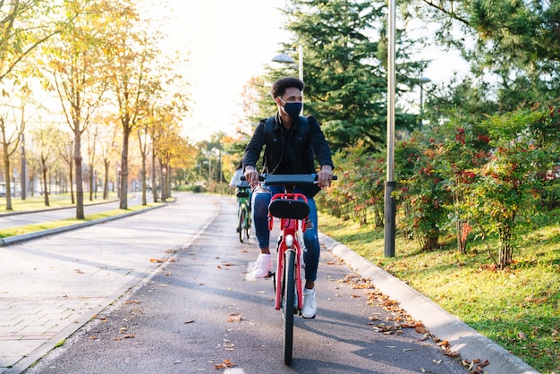 Giovane studente marocchino in sella a una bici elettrica condivisa in un bellissimo parco con molti alberi al tramonto, con indosso una maschera per la pandemia di coronavirus del 2020