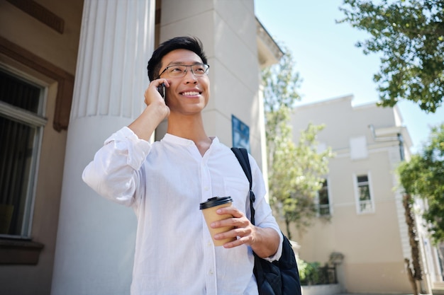 Giovane studente maschio asiatico sorridente attraente in occhiali che parla gioiosamente sul cellulare sulla strada vicino all'università