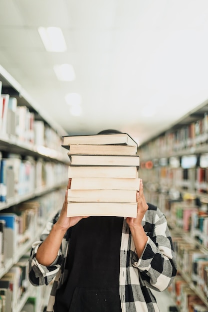 Giovane studente maschio che copre il viso dalla pila di libri al centro della biblioteca del corridoio