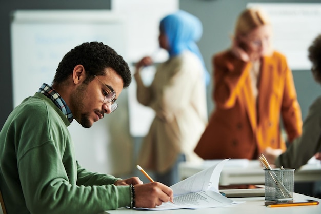 Giovane studente serio in occhiali e pullover casual che prende appunti nel documento