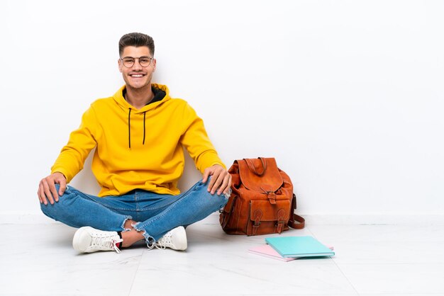 Giovane studente uomo caucasico seduto uno il pavimento isolato su sfondo bianco ridendo