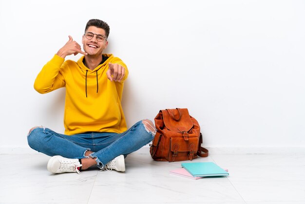 Giovane studente uomo caucasico seduto uno sul pavimento isolato su sfondo bianco che fa il gesto del telefono e indica la parte anteriore