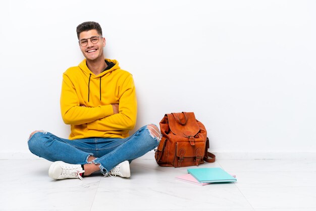 Giovane studente uomo caucasico seduto uno sul pavimento isolato su sfondo bianco con le braccia incrociate e guardando avanti