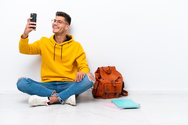 Giovane studente uomo caucasico seduto uno sul pavimento isolato su sfondo bianco facendo un selfie