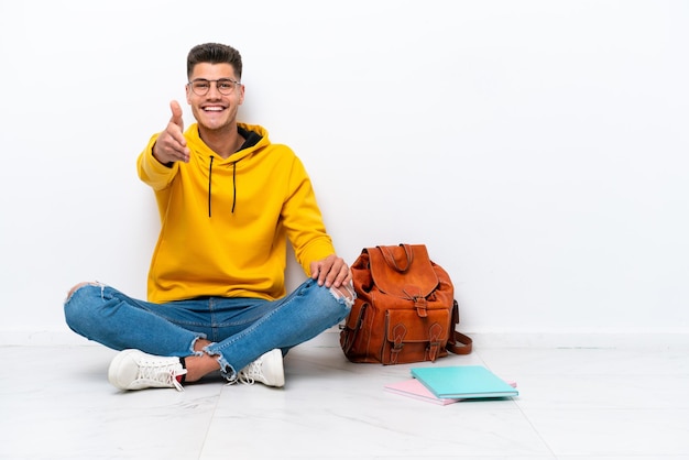 Giovane studente uomo caucasico seduto uno sul pavimento isolato su sfondo bianco stringe la mano per chiudere un buon affare