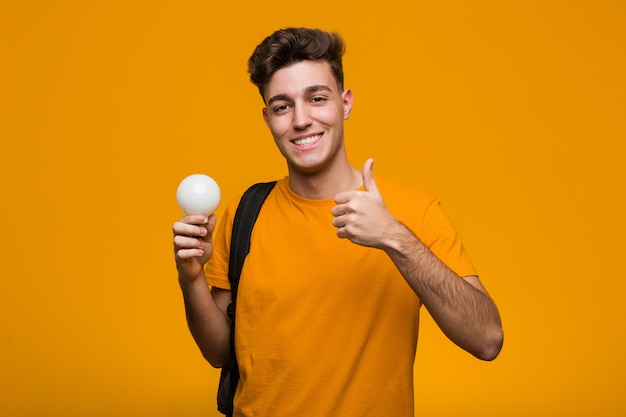 Giovane studente uomo che tiene una lampadina che mostra il segno della vittoria e sorridente ampiamente.