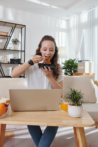 Giovane studentessa abbastanza positiva che mangia noodles per pranzo e guarda la lezione online sul laptop a casa