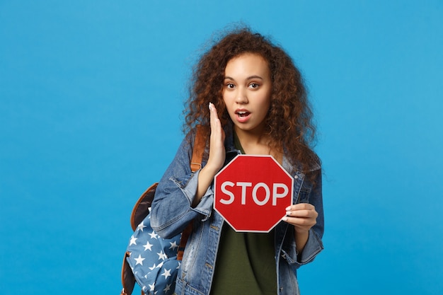 Giovane studentessa adolescente afroamericana in vestiti di jeans, fermata della tenuta dello zaino isolata sulla parete blu