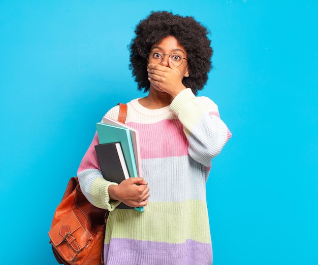 Giovane studentessa afro che copre la bocca con le mani con un'espressione scioccata e sorpresa, mantenendo un segreto o dicendo oops