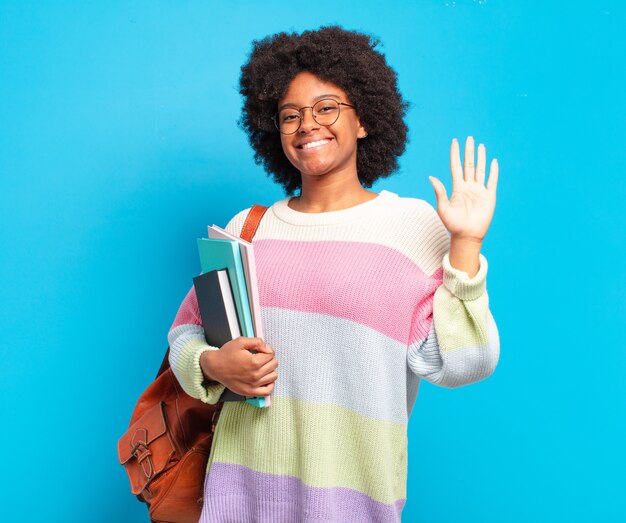 Giovane studentessa afro che sorride allegramente e allegramente, agitando la mano, dandoti il benvenuto e salutandoti o salutandoti