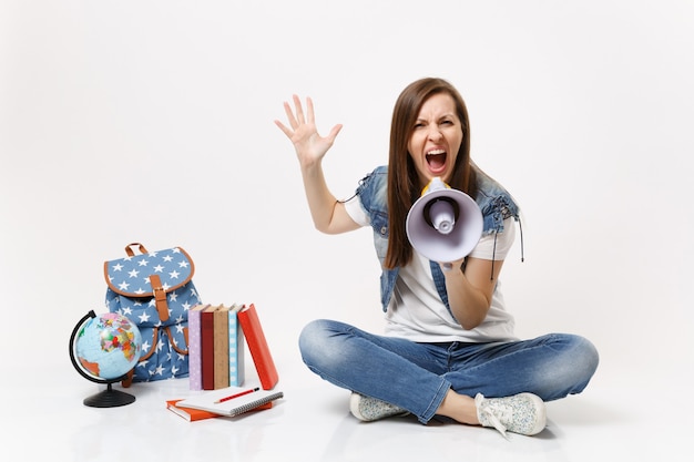 Giovane studentessa arrabbiata irritata urla tenendo il megafono che spande la mano seduto vicino al globo zaino libri scolastici isolati