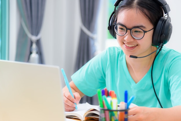 Giovane studentessa asiatica con cuffie e occhiali che scrive sul libro guardando una riunione video su computer portatili una ragazza è felice di prendere la lezione online di apprendimento a distanza di Internet dell'università