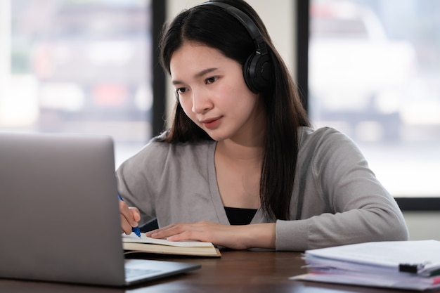 Giovane studentessa asiatica indossa cuffie wireless scrivi sul quaderno per studiare la lingua online guarda e ascolta il docente, webinar tramite videochiamata e-learning a casa, formazione a distanza