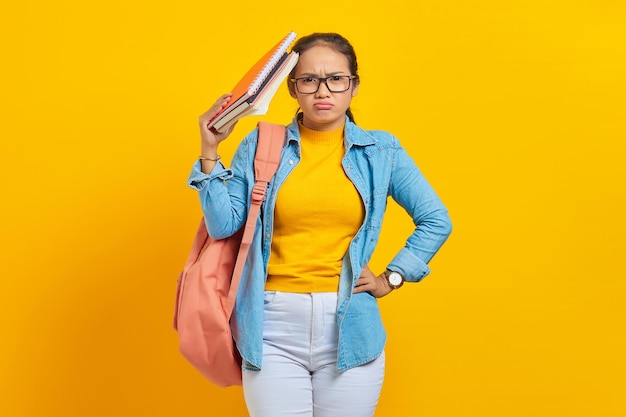 Giovane studentessa asiatica pensierosa in abiti di jeans con zaino in possesso di un taccuino isolato su sfondo giallo Istruzione nel concetto di college universitario di scuola superiore