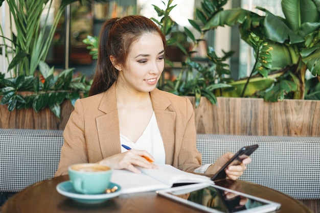 Giovane studentessa asiatica sorridente che fa i compiti in notebook con tablet sul tavolo utilizzando il telefono cellulare al caffè