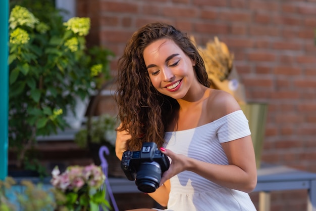 Giovane studentessa attraente o turista che utilizza una fotocamera mirrorless mentre si cammina in città d'estate wo...