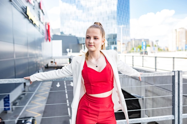 Giovane studentessa bionda in un abito rosso e una giacca bianca in posa nel parcheggio