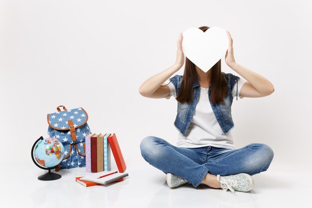 Giovane studentessa bruna che copre il viso con cuore bianco con spazio copia seduto vicino allo zaino del globo, libri scolastici isolati su muro bianco