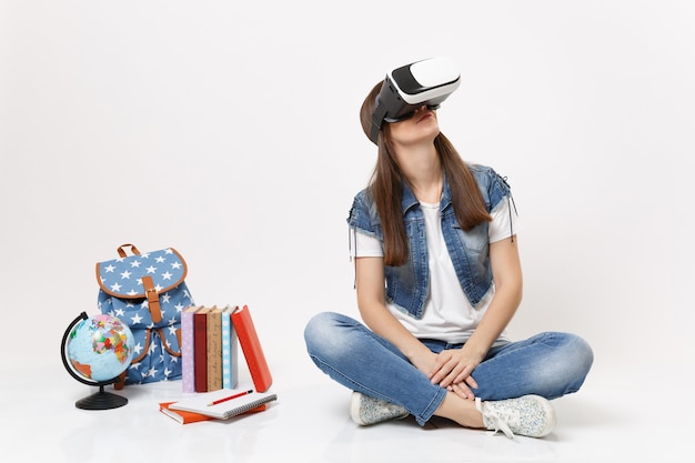 Giovane studentessa bruna con occhiali per realtà virtuale alzando lo sguardo godendosi seduta vicino al globo, zaino, libri scolastici isolati su muro bianco