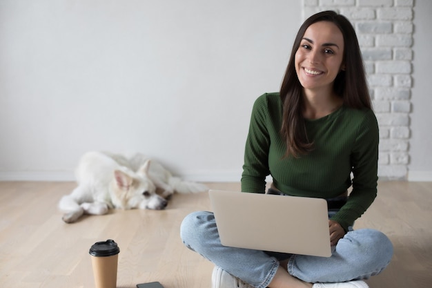 Giovane studentessa carina e sicura di sé in abiti casual sta utilizzando un computer portatile e sta sorridendo mentre era seduta sul pavimento su sfondo grigio Concetto di laureato dell'università di istruzione di studio