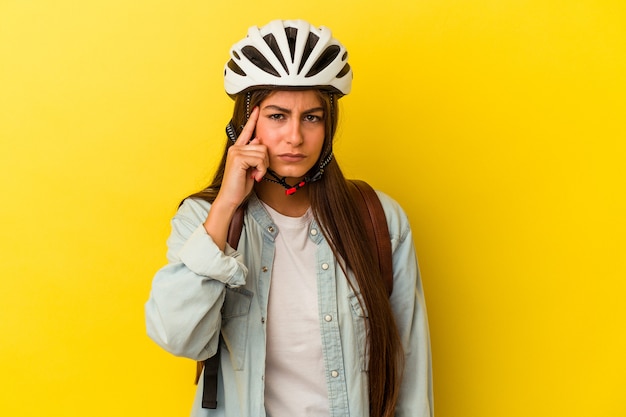 Giovane studentessa caucasica che indossa un casco da bici isolato su sfondo giallo che punta il tempio con il dito, pensando, concentrato su un compito.