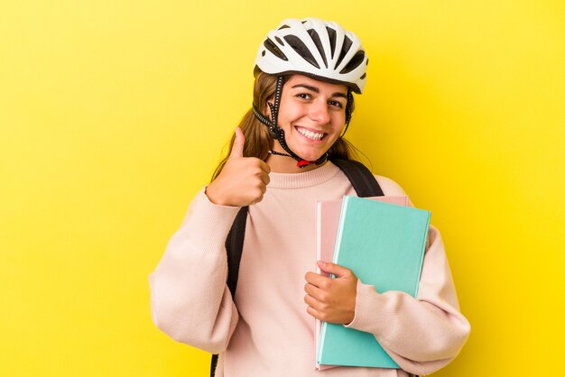 Giovane studentessa caucasica che indossa un casco da bici isolato su sfondo giallo sorridente e alzando il pollice