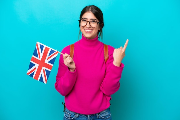 Giovane studentessa caucasica che tiene bandiera inglese isolata su sfondo blu che mostra e alza un dito in segno di meglio