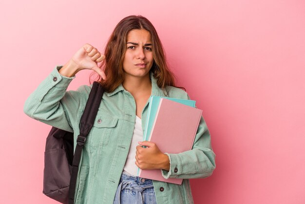 Giovane studentessa caucasica che tiene in mano libri isolati su sfondo rosa che mostra un gesto di antipatia, pollice in giù. Concetto di disaccordo.