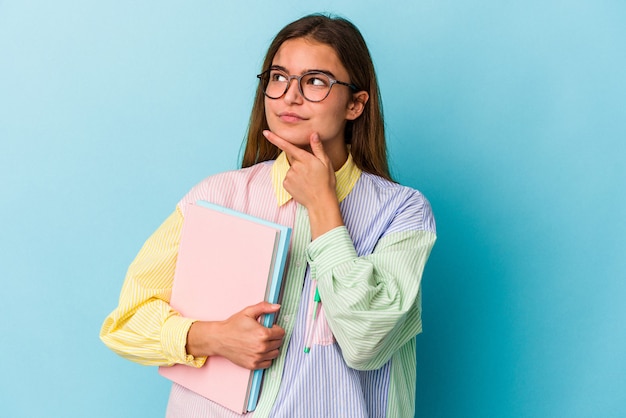 Giovane studentessa caucasica che tiene libri isolati su sfondo blu guardando di traverso con espressione dubbiosa e scettica.