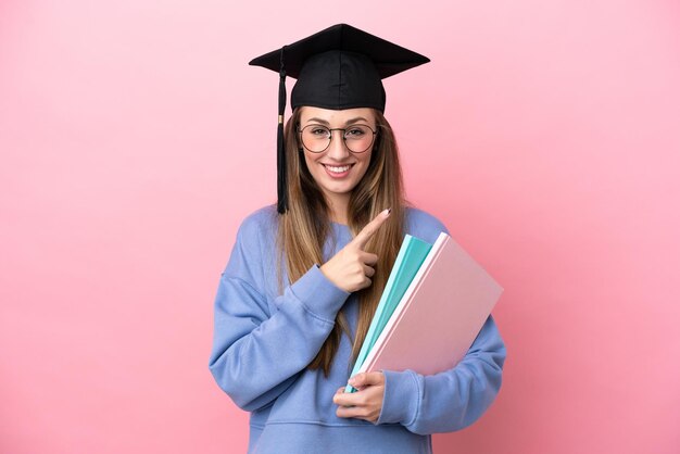 Giovane studentessa che indossa un cappello laureato isolato su sfondo rosa che punta al lato per presentare un prodotto