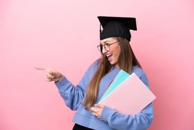 Giovane studentessa che indossa un cappello laureato isolato su sfondo rosa che punta il dito di lato e presenta un prodotto