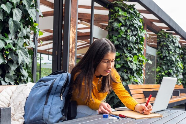 Giovane studentessa che scrive sul suo taccuino accanto al suo computer portatile seduto a un tavolo fuori