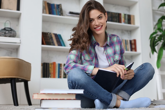 Giovane studentessa che studia a casa, scrive articoli per la pubblicazione, seduta sul pavimento contro un accogliente interno domestico, circondata da una pila di libri.