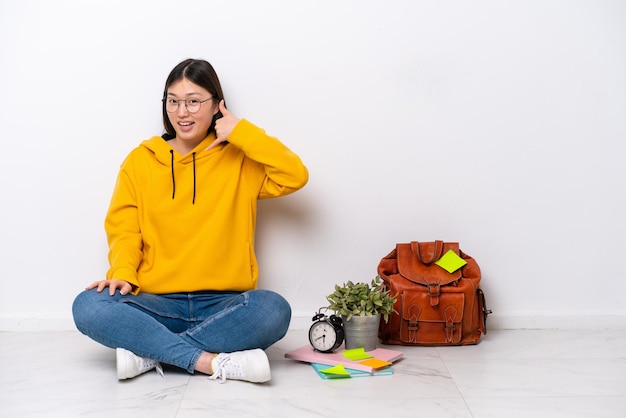 Giovane studentessa cinese seduta sul pavimento isolata sul muro bianco che fa il gesto del telefono Chiamami indietro segno