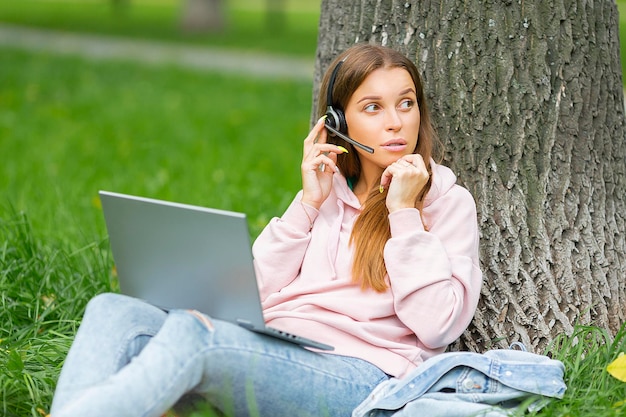 Giovane studentessa con auricolare e laptop seduto sull'erba nel parco del campus