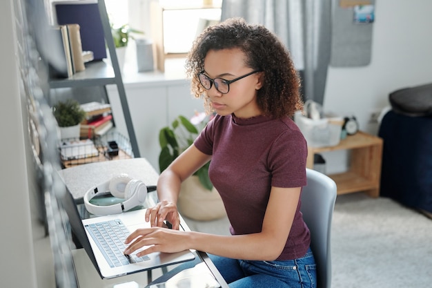 Giovane studentessa contemporanea in abbigliamento casual seduta da un tavolino davanti al laptop mentre segue un corso di studio online in camera da letto