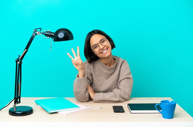 Giovane studentessa di razza mista che studia su un tavolo felice e conta tre con le dita