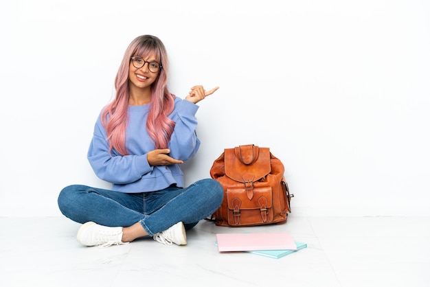 Giovane studentessa di razza mista con i capelli rosa seduta su un pavimento isolato su sfondo bianco che estende le mani di lato per invitare a venire